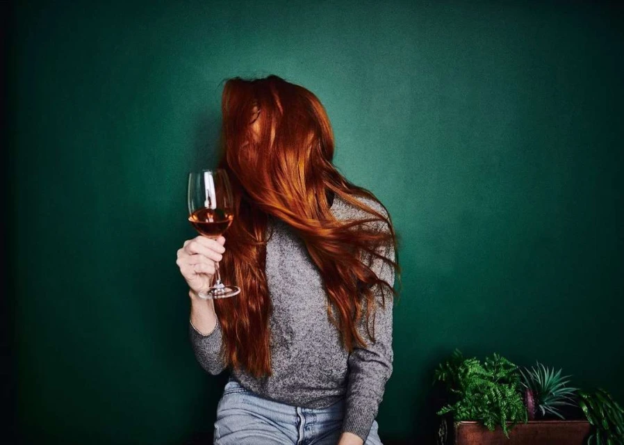 A woman with long red hair holding a wine glass against a dark green background, wearing a gray sweater and jeans.