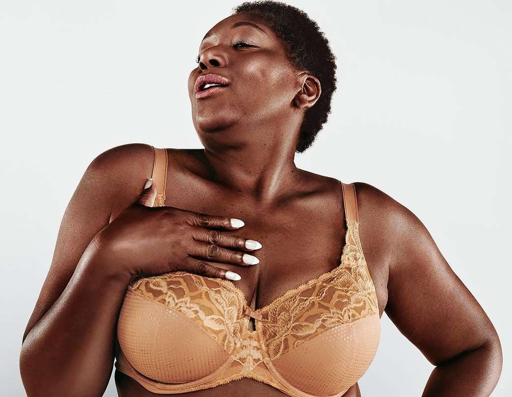 An African-American woman poses in a bra against a white background. Her hand rests on her chest, eyes closed, as the light illuminates her skin.