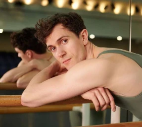 A young man leaning on a wooden ballet barre in a dance studio, reflected in a mirror behind him