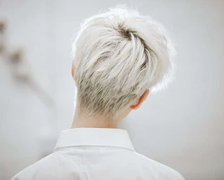 Close-up back view of a person with short white platinum hair wearing a white collared shirt against a pale background