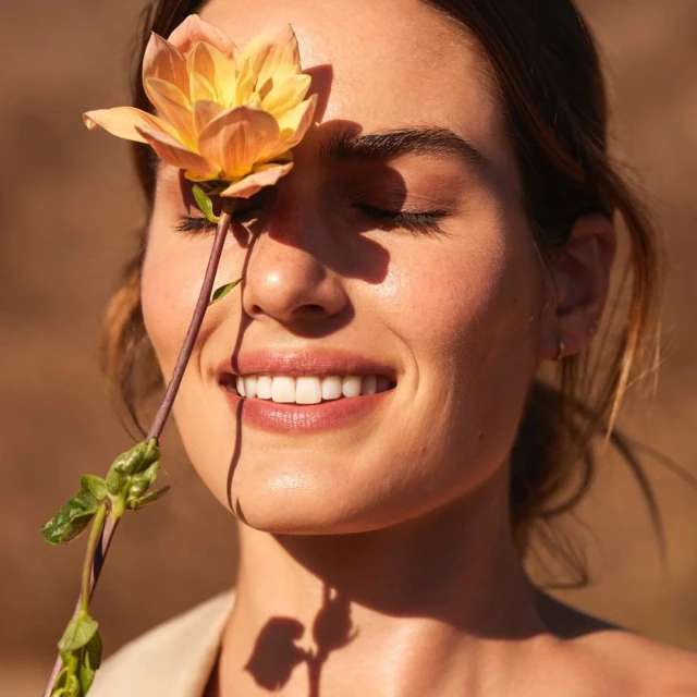 Close-up of a woman with eyes closed, a gentle smile, and a yellow flower over one eye in a natural setting with soft, diffused light.