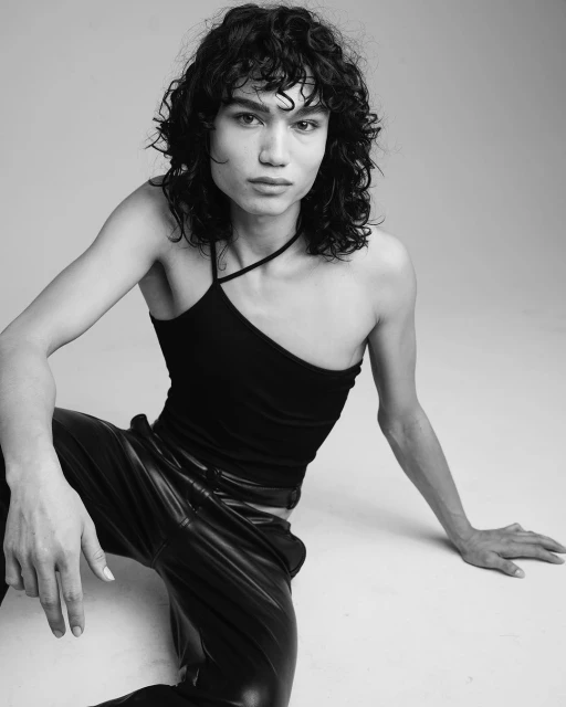Black and white portrait of a person with curly hair, wearing a one-shoulder top and leather pants, posing on a neutral backdrop.