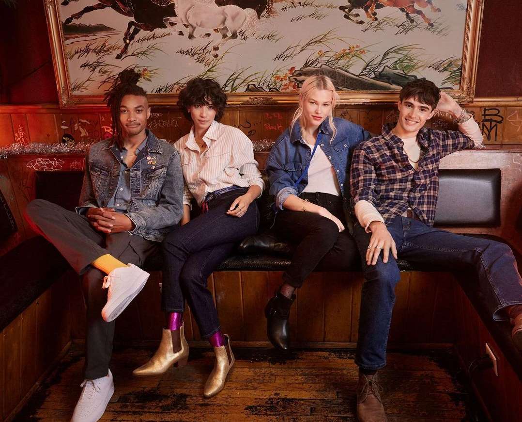 Group of four young adults sit in a vintage booth. Two women and two men pose with denim, casual shirts, and boots, indoors.