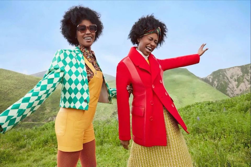 Two women pose outdoors in bright 70s fashion: patterned blazers, yellow overalls, hosiery, and dresses against a green hillside backdrop.
