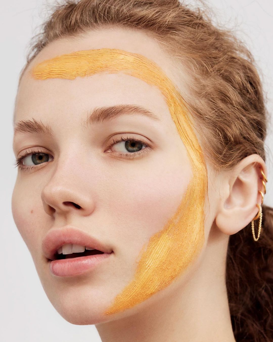 Close-up shot of a young woman with golden face paint, fair skin, and curly hair against a plain background in a beauty context.