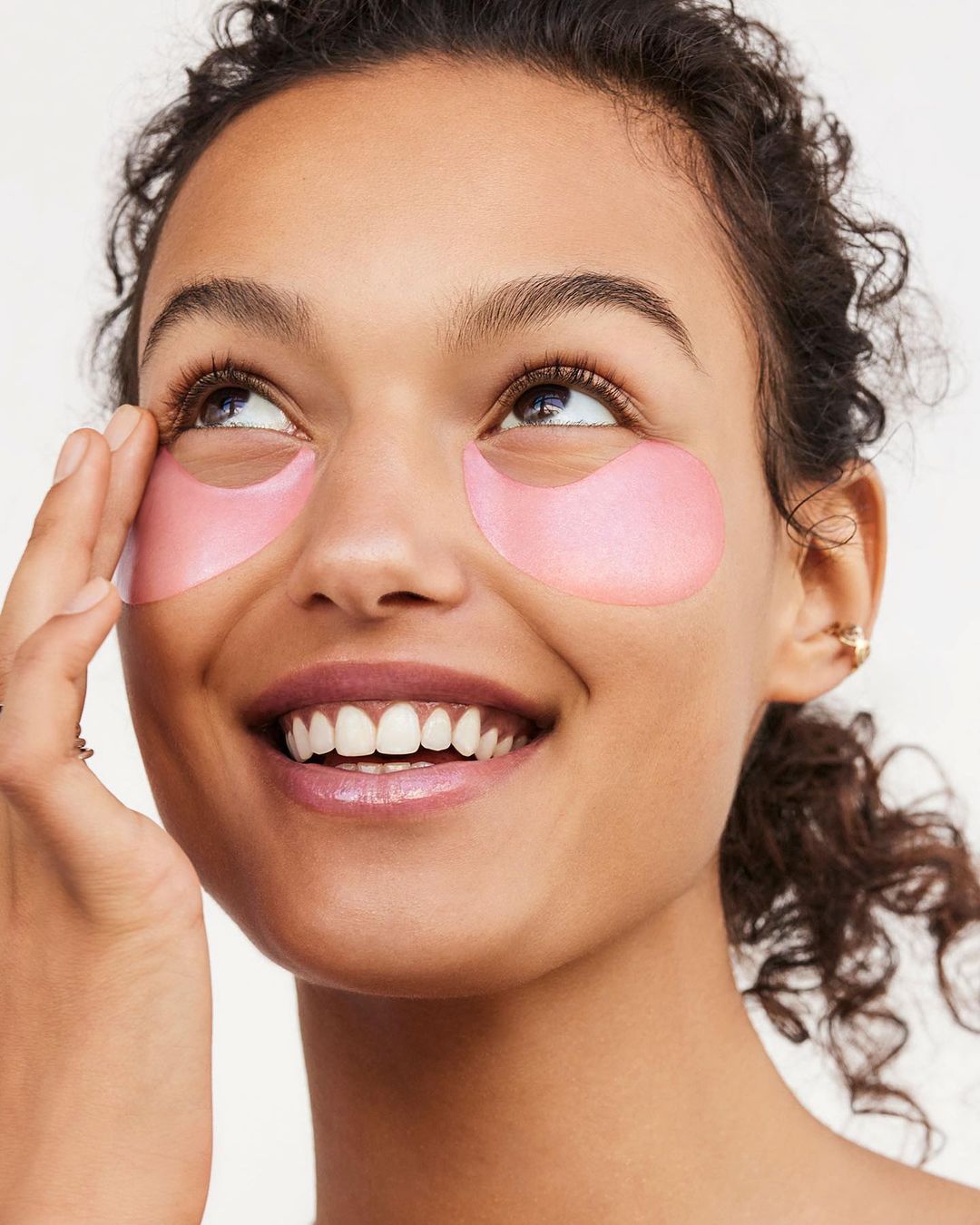 Close-up of a young woman with pink under-eye patches smiling and looking upward, demonstrating skincare in a bright, studio setting.