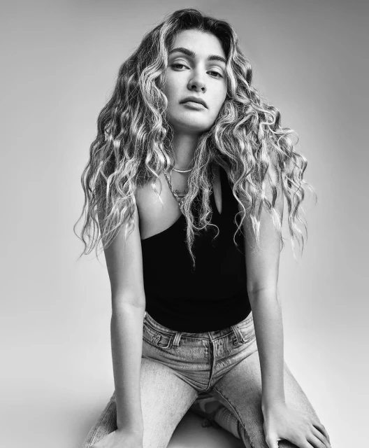 Portrait of a young adult woman with curly hair, wearing a black top and jeans, posing in a photo studio with neutral background.