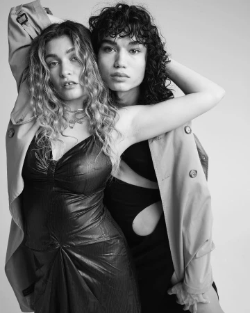Two models posing closely together wearing black dresses and a shared tan trench coat against a plain background.
