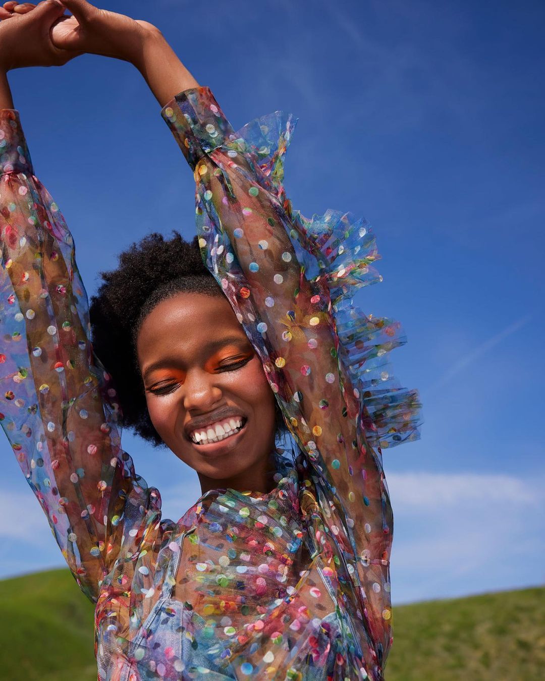 Smiling woman in polka dot dress against a blue sky and green field. Fashion portrait with bright makeup and cheerful pose in nature.