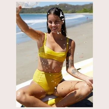 A woman in a yellow bikini sits on a surfboard at the beach, pouring water over her head with the ocean and sky in the background.