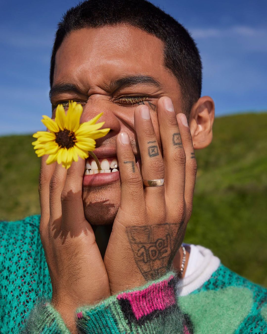 Close-up of an adult with a yellow flower between their teeth, hands on face, wearing a patterned sweater outdoors on a grassy hill.