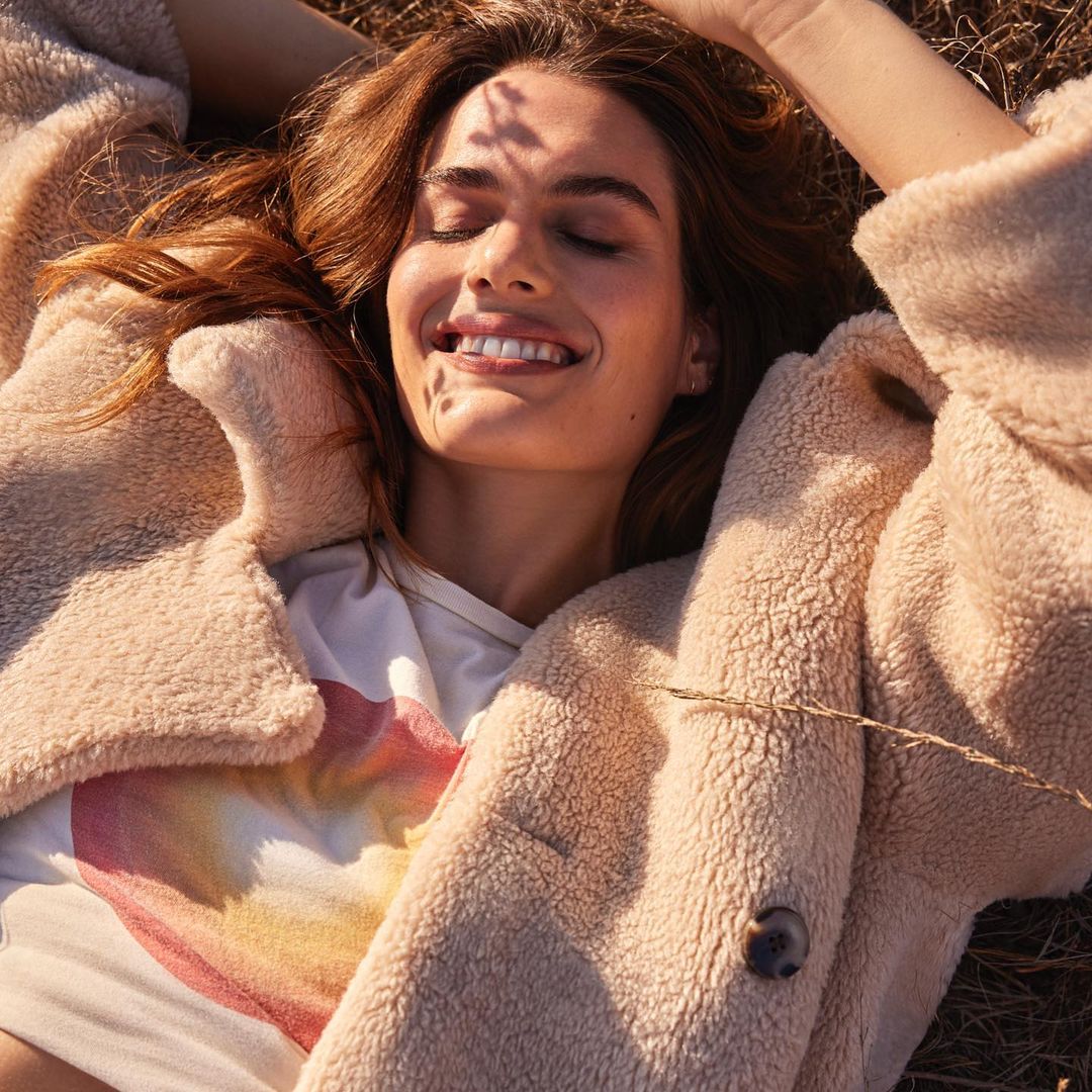Attractive young woman with brown hair lying in a field of dry grass, wearing a teddy bear jacket over a shirt with a heart graphic.