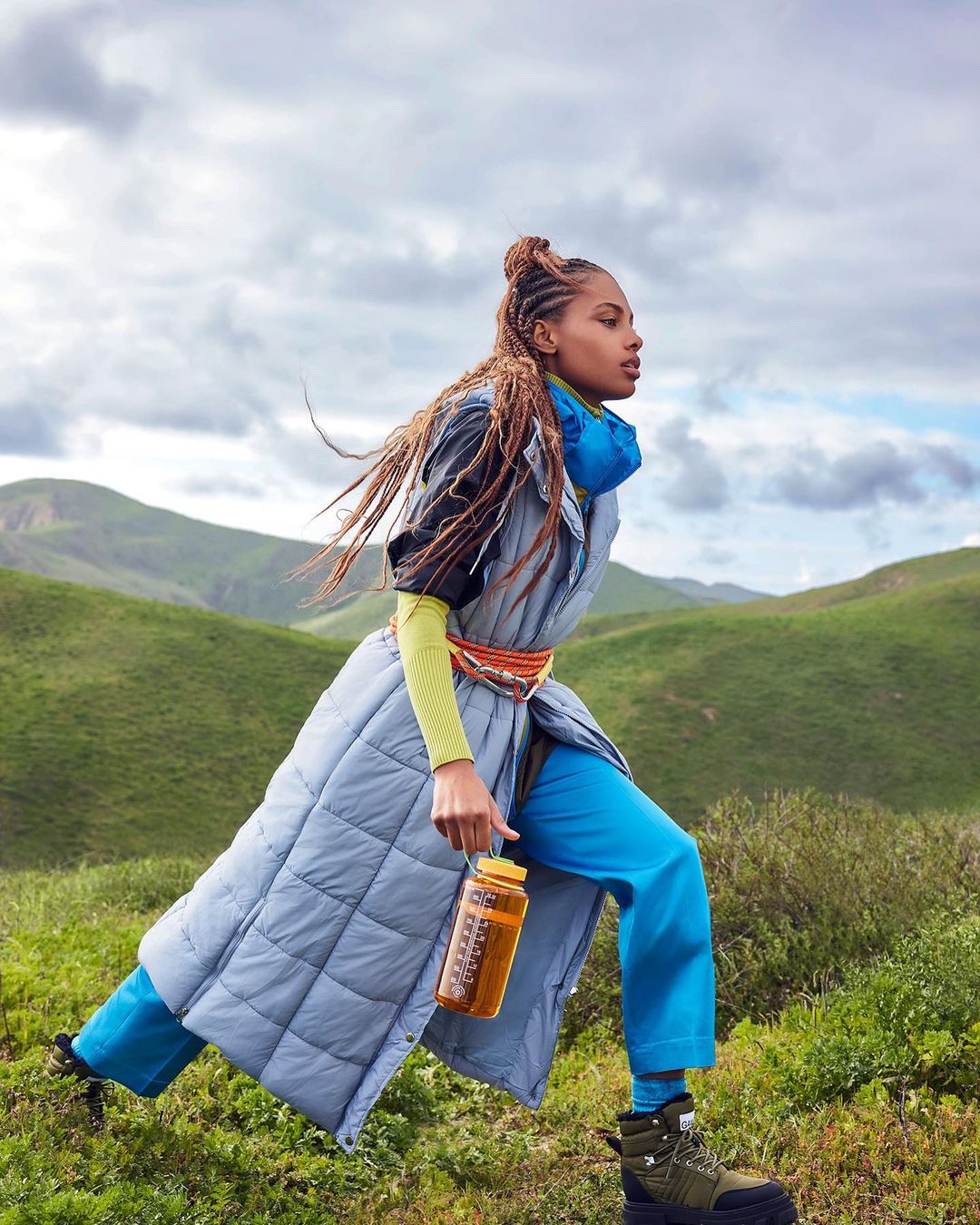 Woman in athleisure wear strides confidently across a grassy hillside, carrying a water bottle, with a backdrop of rolling green hills.