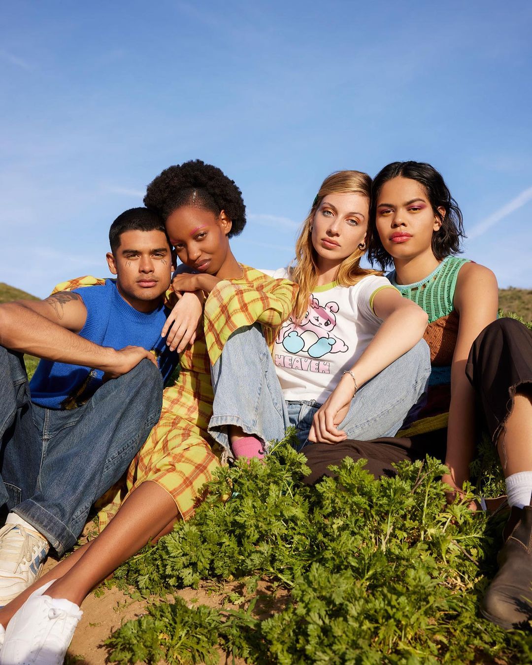 Four young adults, two women and two men, are seated together on a grassy hill under a blue sky, showcasing casual fashion and relaxed poses.