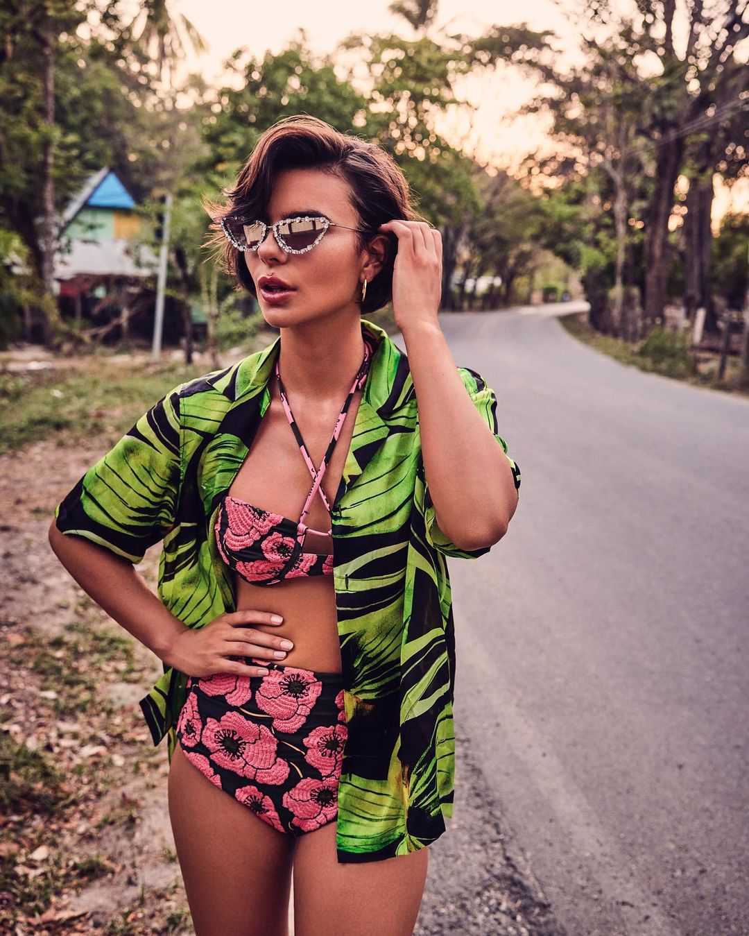 Brunette woman in floral bikini and leaf-print shirt poses on a tropical road with palm trees and a distant house.