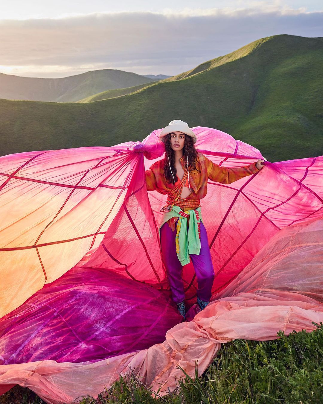 Woman stands amid pink parachute on a grassy hill with green mountains in the background, wearing purple pants, shirt and white hat.