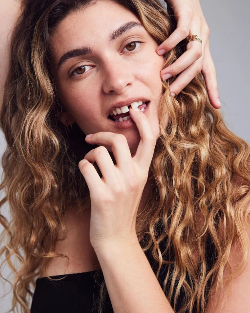 Young woman with long wavy hair and a missing front tooth poses in a studio setting with another hand gently touching her face.
