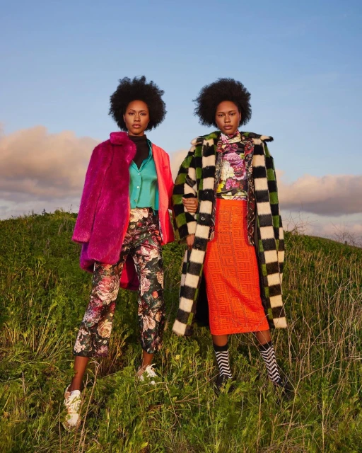 Two Black women wearing brightly-colored coats and patterned pants stand on a grassy hill under a blue sky with scattered clouds.