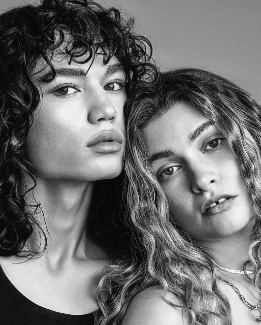Black and white portrait of two women with curly hair, head and shoulders, looking at the camera, with a neutral studio background.