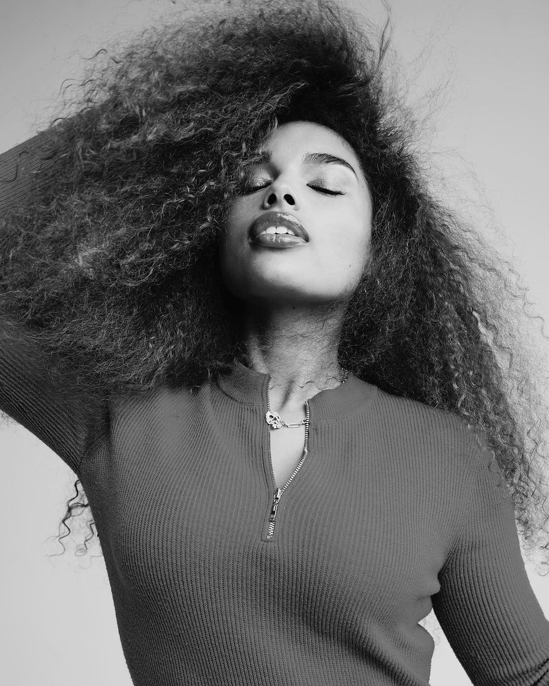 Black and white portrait of a woman with curly hair and closed eyes, wearing a ribbed top with a zipper and a delicate necklace.
