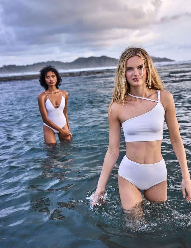Two women in white swimsuits stand in ocean water; overcast sky and rocky shoreline in the background. Fashion and beauty shot.