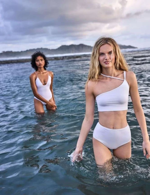 Two women in white swimsuits stand in ocean water; overcast sky and rocky shoreline in the background. Fashion and beauty shot.