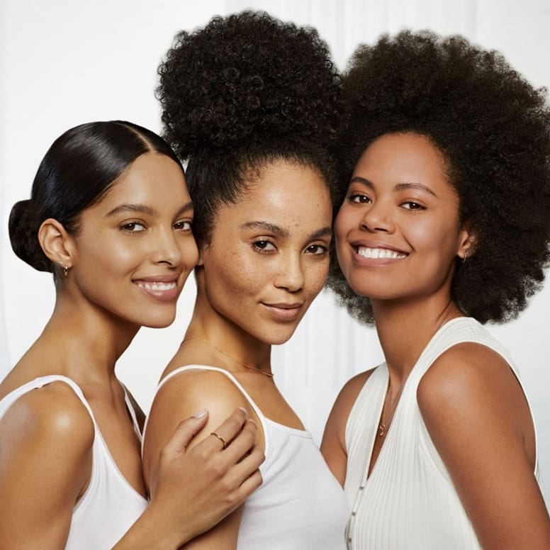 Three women with diverse hairstyles pose closely together. Their natural hair and radiant skin are highlighted in the close-up studio portrait.