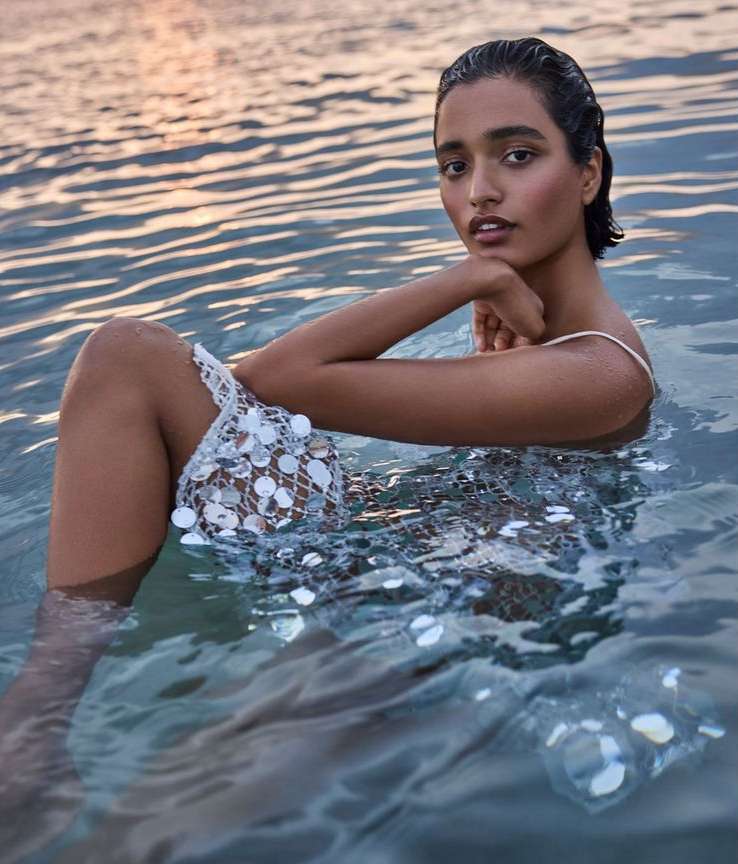 A woman wearing a white beaded dress sits in shallow water as the sun sets in the background. She looks at the camera.