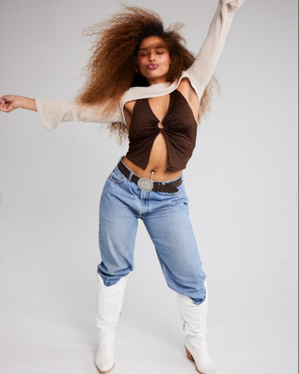 A young woman with curly hair poses in a brown top, jeans, and white boots against a white backdrop in a studio setting.
