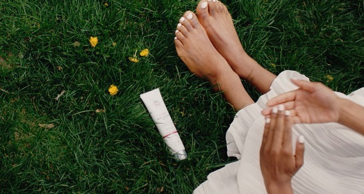 Woman applies lotion outdoors while sitting on grass, featuring a white tube of beauty product for skincare or self-care.