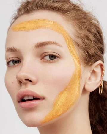Close-up portrait of a woman with a wide streak of orange cosmetic pigment swiped across her forehead and cheek by Sarah Kehoe.