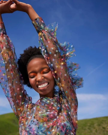 Woman in sheer polka dot top with arms raised against clear blue sky and green hills