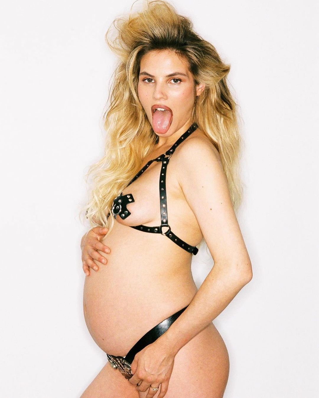 Pregnant woman in black lingerie with long blonde hair poses against a white backdrop with tongue sticking out, hands on belly.