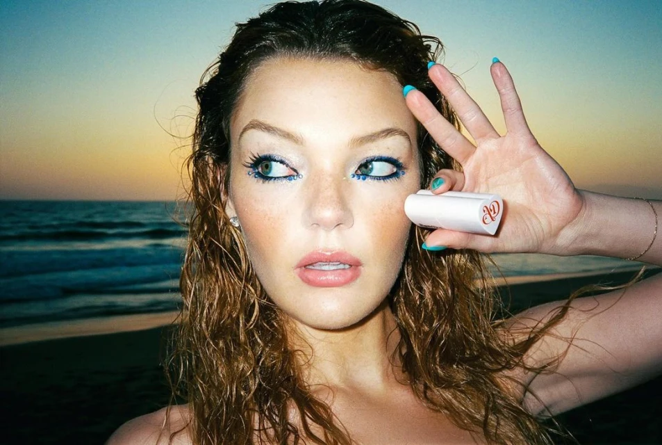 Close-up of a woman with glittery blue eye makeup posing on a beach at sunset, holding a small tube of white beauty product.