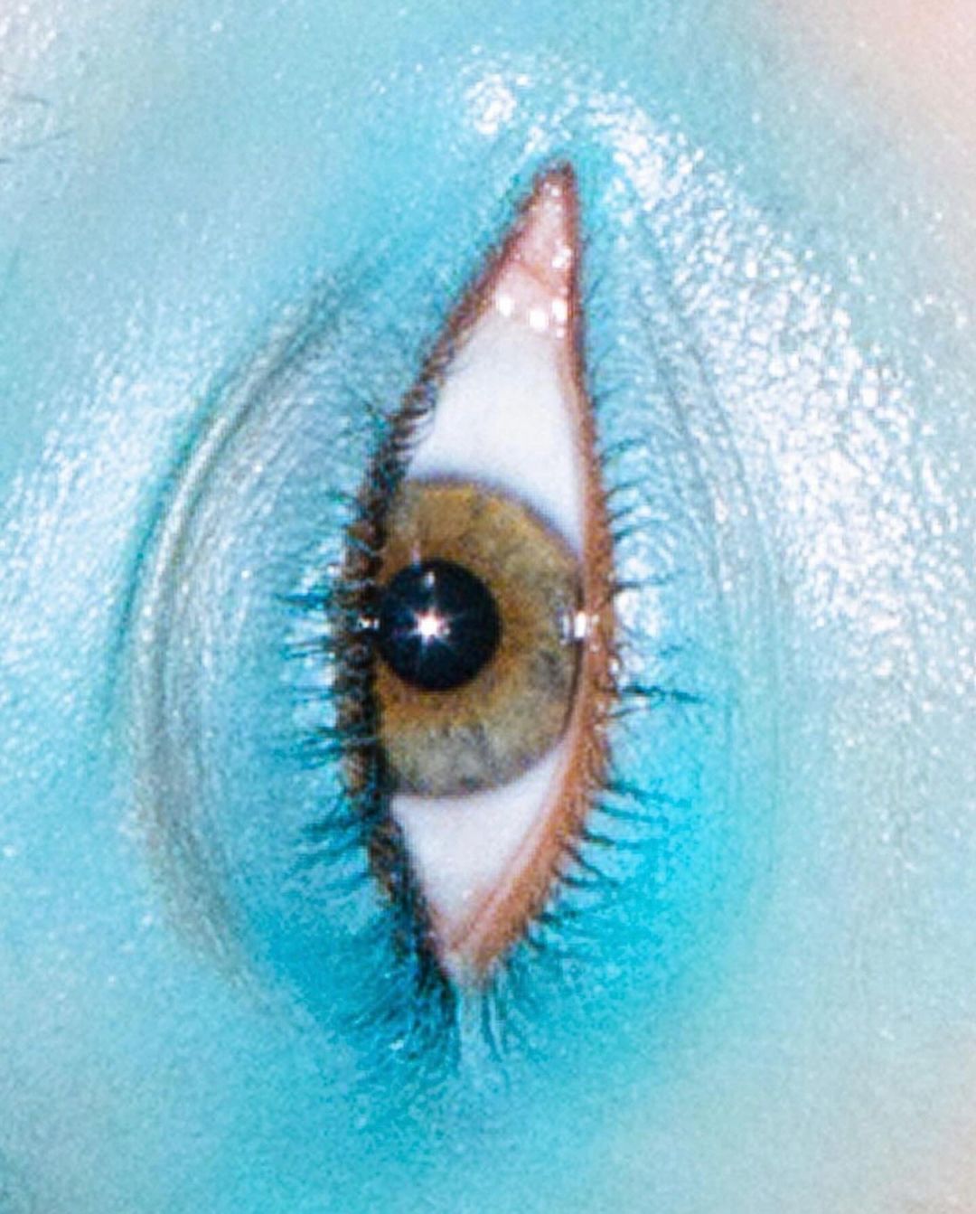 Extreme close-up of a human eye with turquoise blue skin surrounding it. The eye has a brown iris and long lashes.