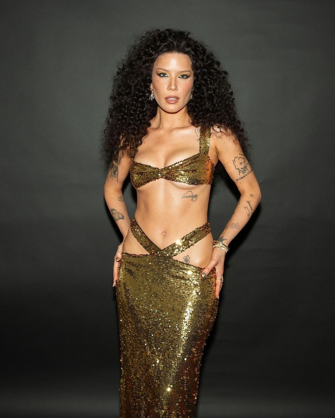 Halsey in a gold sequin bra and skirt posing against a dark gray backdrop, showcasing her tattoos and long curly hair.