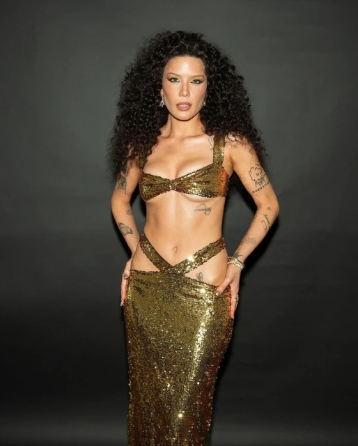 Halsey in a gold sequin bra and skirt posing against a dark gray backdrop, showcasing her tattoos and long curly hair.