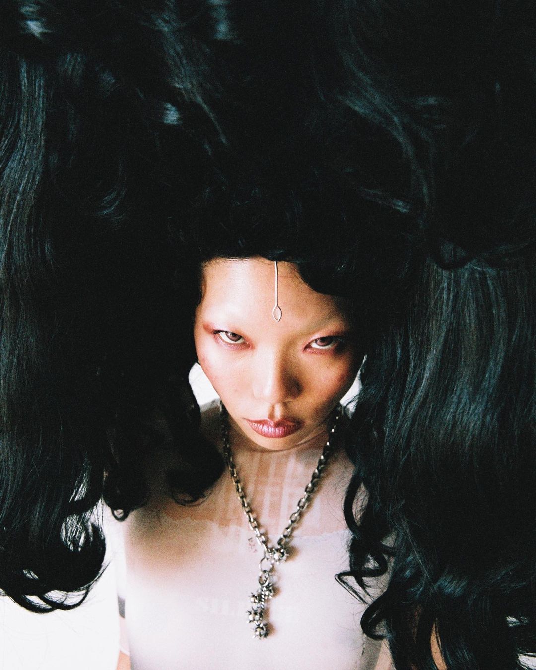 Woman with dramatic black hair styled in a high upward fashion, pale complexion, wearing a delicate silver chain and modern jewelry.