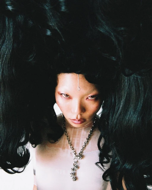 Woman with dramatic black hair styled in a high upward fashion, pale complexion, wearing a delicate silver chain and modern jewelry.