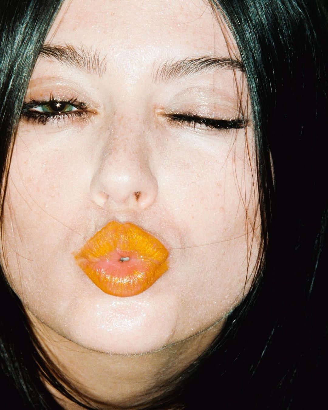 Close-up portrait of an adult female with dark hair winking, pouty lips covered in bright orange lipstick, and fair skin.
