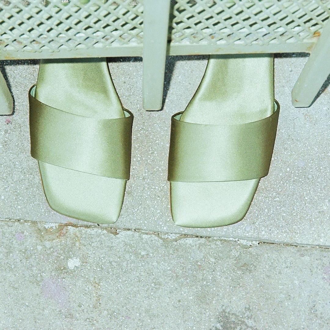 Close-up of olive-green satin platform heels on a gray concrete surface, partially obscured by a pale-green metal chair.