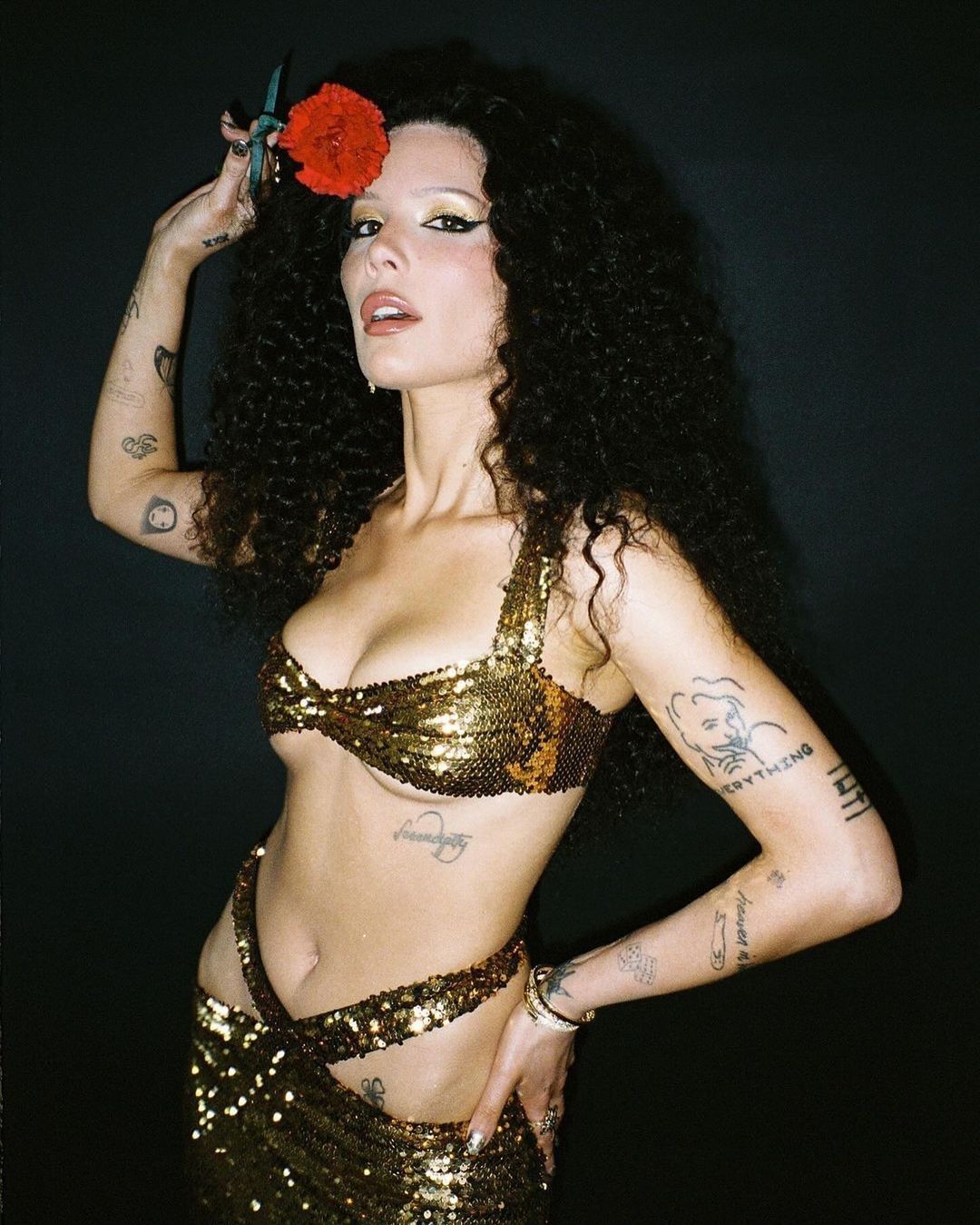 Glamorous portrait of a woman with curly hair, wearing a sequin outfit, holding a red flower, in a dimly lit indoor setting.