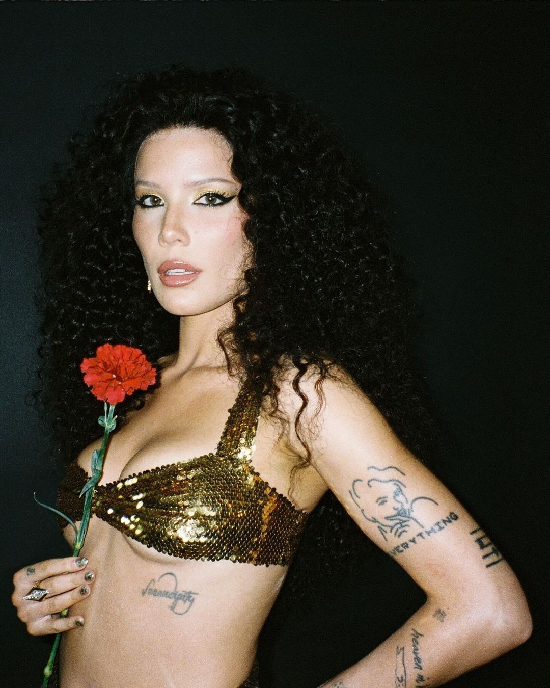 Portrait of a woman with curly hair, wearing a gold sequin top, holding a red flower, with a dark background.