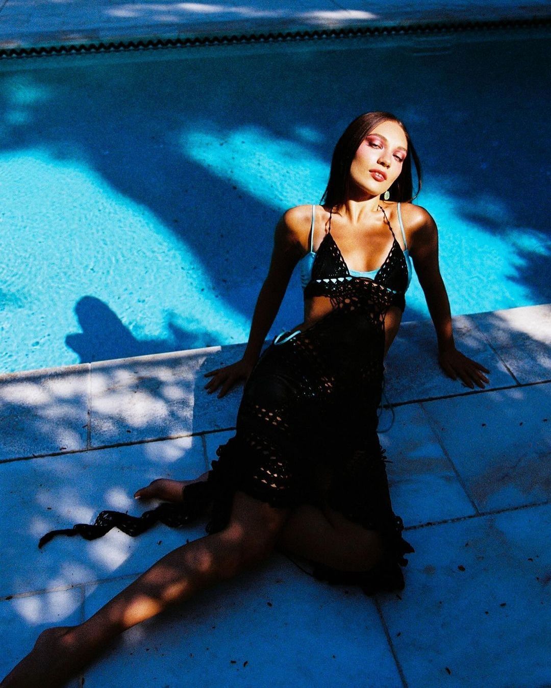 A woman in a black crocheted dress poses by a bright blue swimming pool on a sunny day, highlighting fashion and outdoor lifestyle.