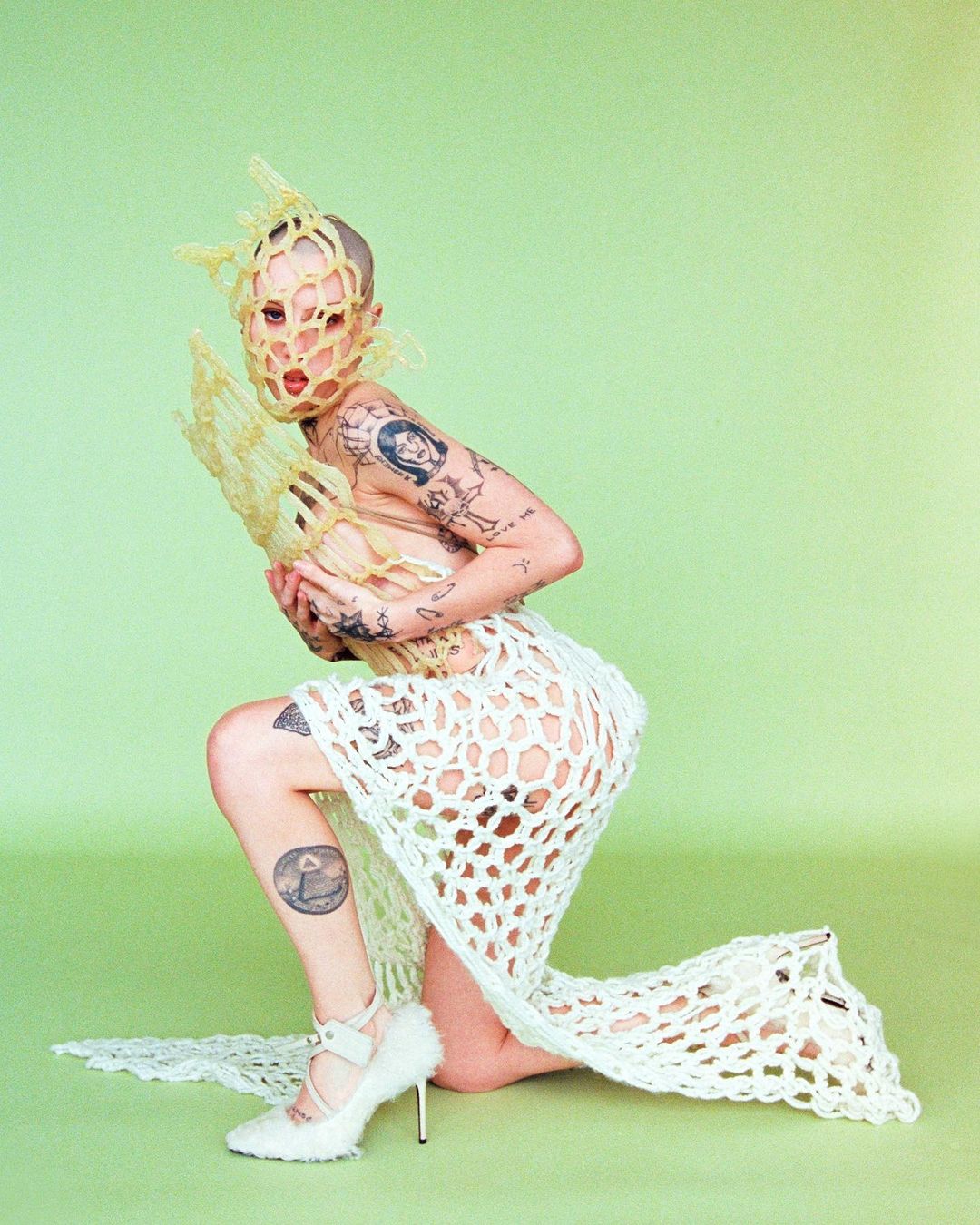 Conceptual fashion photo of a bald woman in a netted mask and dress kneeling in front of a light green backdrop with neutral color grading.