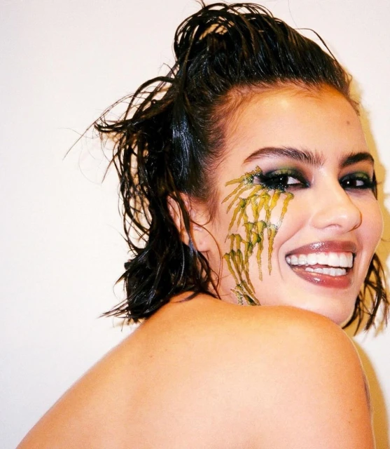 Smiling woman with wet hair and green makeup in a close-up shot against a bright white background, fashion beauty portrait.