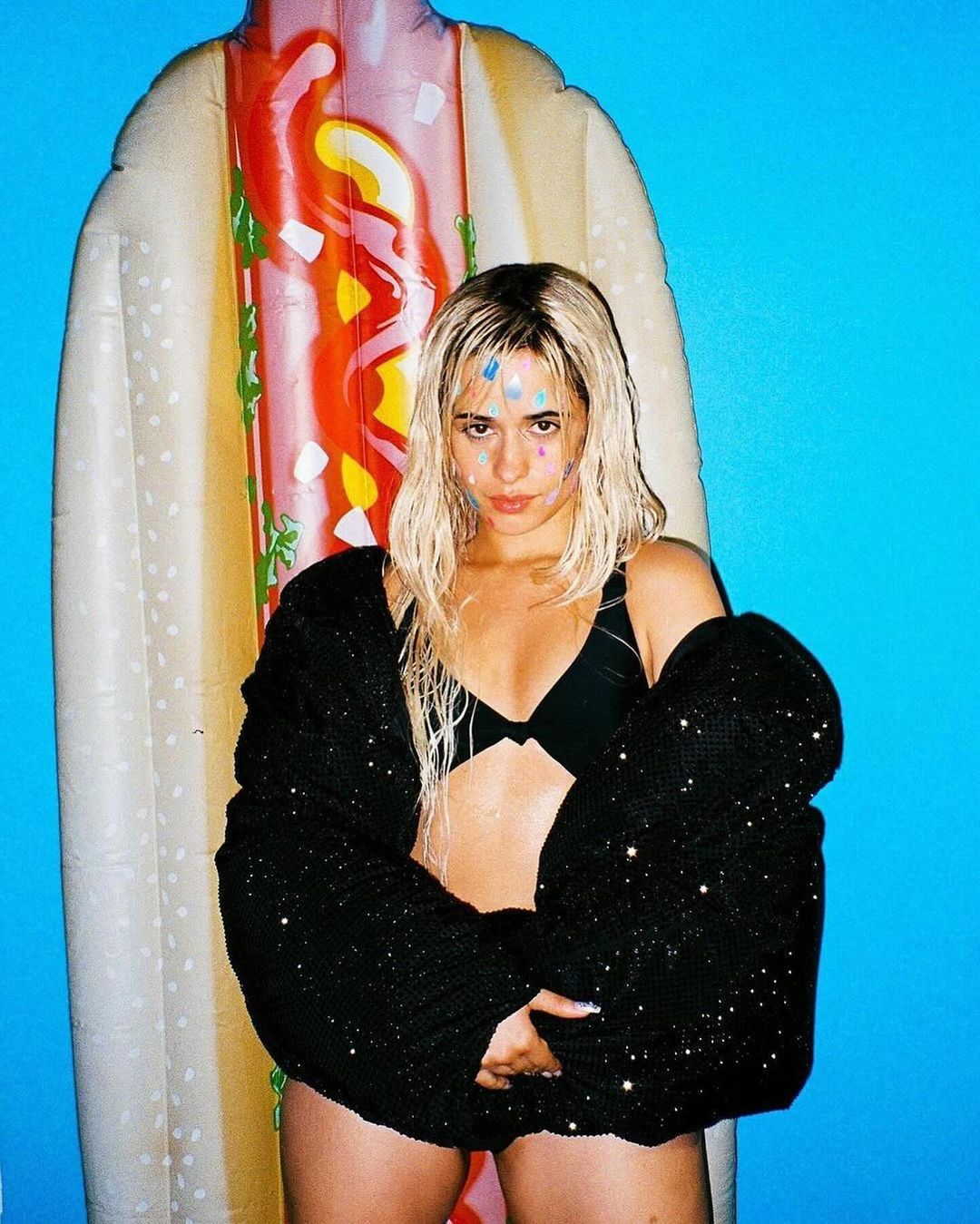 A woman poses in a black bikini top and sparkling cardigan in front of an inflatable hot dog against a bright blue backdrop.