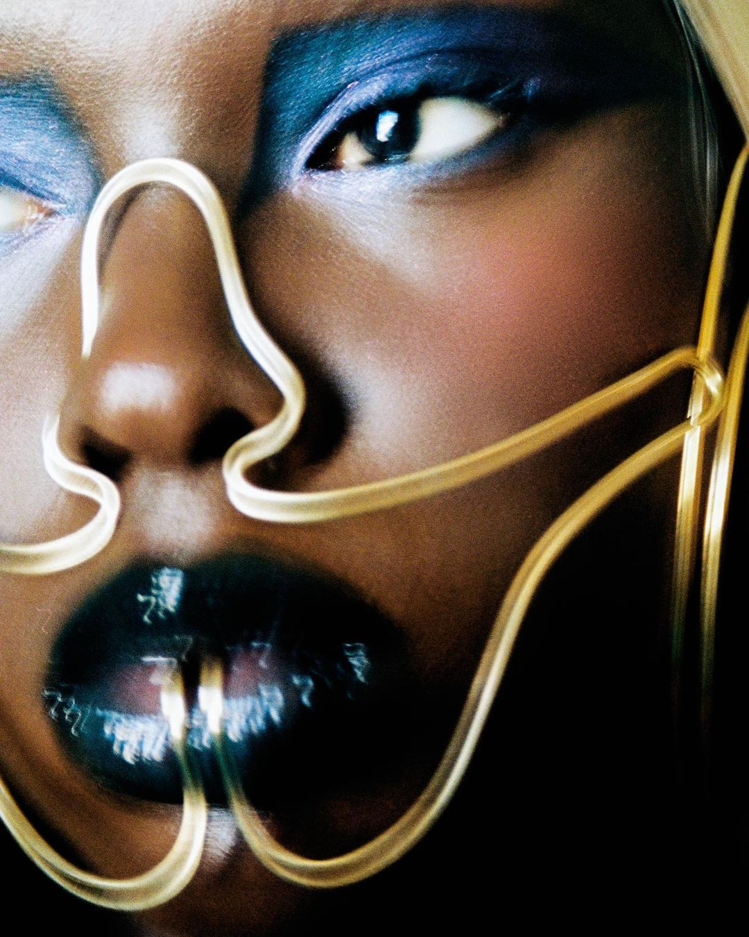 Close-up portrait of a Black woman with dramatic blue eyeshadow and glossy lips, face framed by gold wire art.