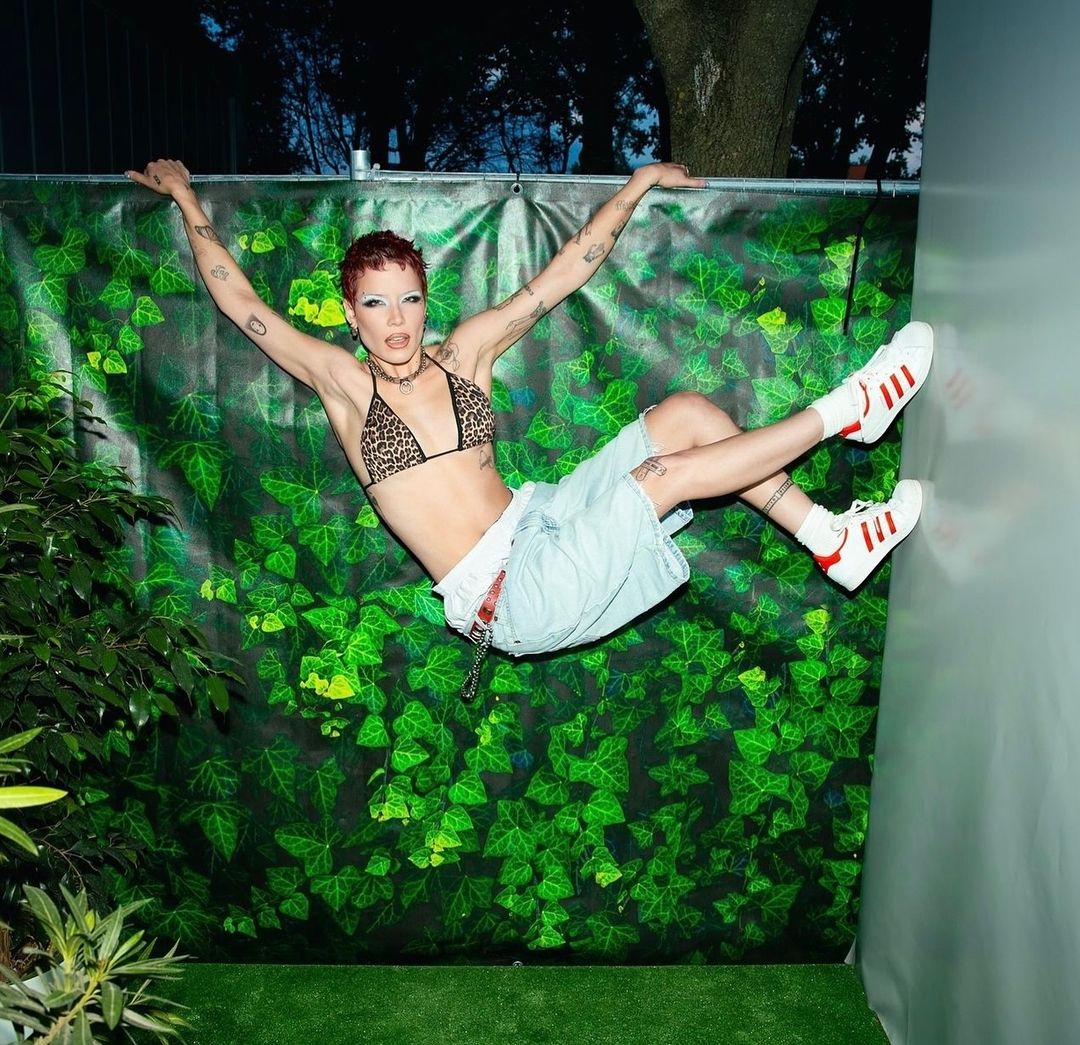 Woman poses with arms raised wearing a leopard-print bikini top, shorts, and sneakers against a green ivy background in a studio.