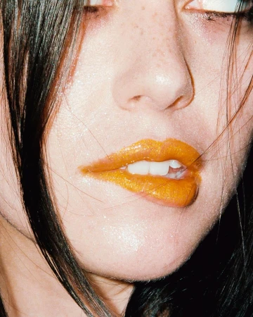 Close-up of a young woman's face showcasing gold lipstick and dark hair, emphasizing beauty and makeup in an indoor setting.